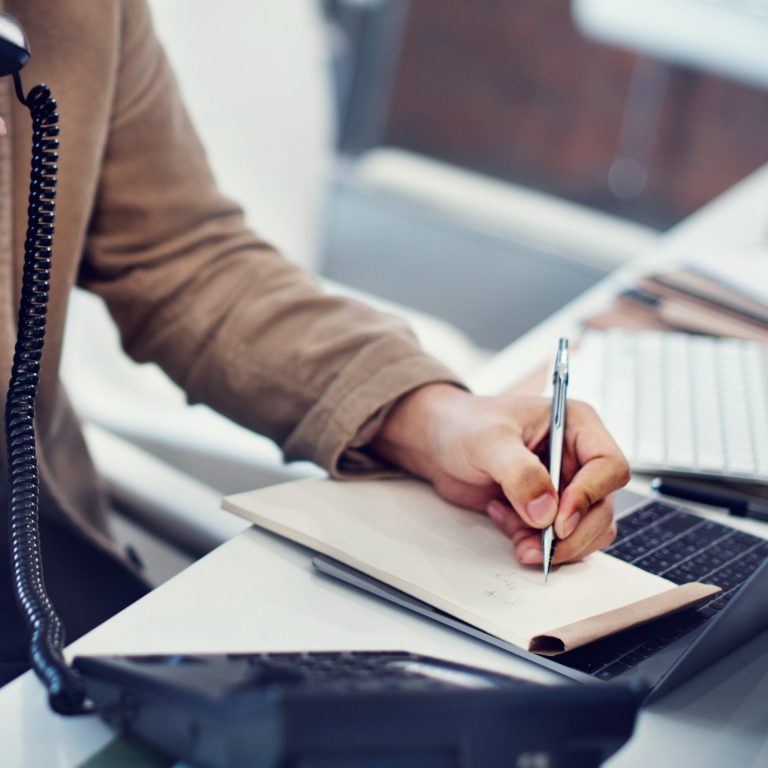 Eine Hand schreibt auf einem Notizblock, während ein Laptop und Telefon sichtbar sind.