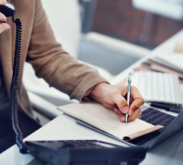 Person am Schreibtisch telefoniert und macht Notizen in ein Notizbuch.